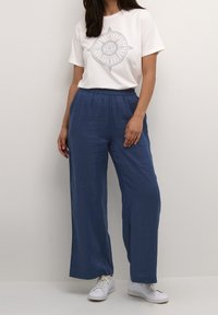 White t-shirt with circular graphic design, paired with loose, navy blue linen pants. Model wears white sneakers. Simple, casual look.