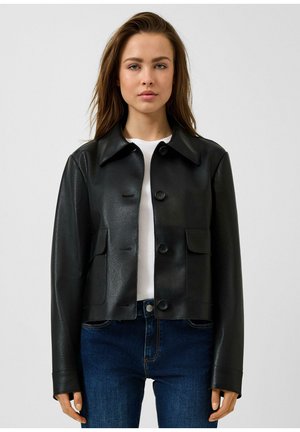 Frau trägt eine schwarze, geknöpfte Kunstlederjacke über einem weißen Hemd und blauen Jeans, steht vor einem einfarbigen hellen Hintergrund.