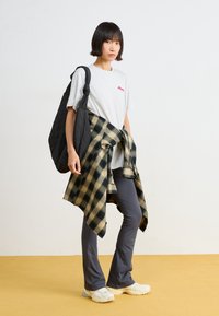 Camiseta gris con logo rojo, bolso acolchado negro, camisa a cuadros verde y beige atada a la cintura, pantalones ajustados grises, zapatillas blancas.