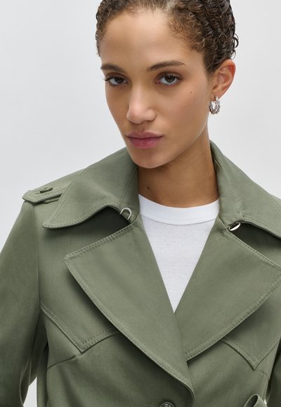 Γυναίκα με κοντά κατσαρά μαλλιά που φοράει ασημένια κρίκους σκουλαρίκια και χακί πράσινο παλτό τύπου τρεντσκότ πάνω από λευκή ριμπ μπλούζα, μπροστά σε ένα απλό φόντο.