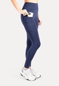 Marineblauwe legging met hoge taille en zijzak, rekbare stof, gladde textuur, gecombineerd met witte en zilveren sportschoenen.