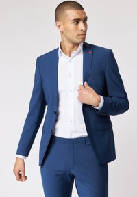 Giacca blu con una texture sottile, abbinata a una camicia bianca. Presenta linee eleganti, due bottoni e un accento rosso sul rever.