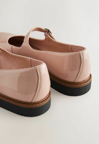 Chaussures Mary Jane brillantes couleur nude avec une bride sur le dessus du pied, semelles intermédiaires marron et semelles extérieures épaisses en caoutchouc noir.