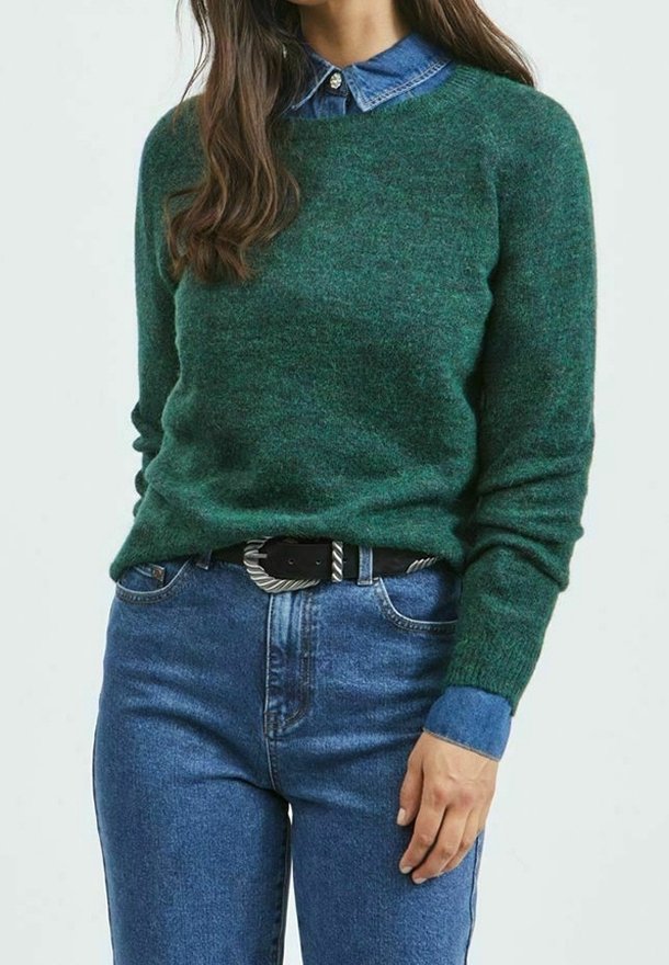 Suéter de lana verde con mangas largas, escote redondo y una textura suave, llevado sobre una camisa de mezclilla con cuello y combinado con jeans azules.
