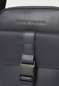 Navy blue shoulder bag made of textured synthetic material, featuring a prominent clasp closure and the brand name "Tommy Hilfiger" embossed.