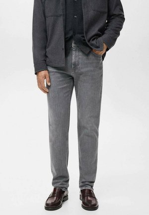 Man wearing grey jeans, dark button-up shirt, and dark brown leather loafers, standing with one hand in pocket.