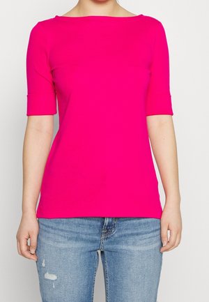 Femme portant un haut rose vif à manches courtes et un jean bleu clair déchiré, debout devant un fond blanc uni.