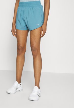 Person iført blå Nike løbeshorts og hvide Nike sneakers står mod en ensfarvet hvid baggrund.