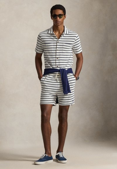 Fehér és tengerészkék csíkos rövid ujjú ing, elöl gombolós, hozzá illő csíkos rövidnadrággal és derekán megkötött tengerészkék pulóverrel.