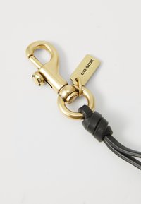 Gold metal keychain clip with a black leather strap, featuring a round ring and a tag embossed with "COACH." Smooth finish and minimal design.