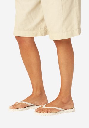 Ben iført beige shorts og off-white flip-flops med teksturerede såler på hvid baggrund.