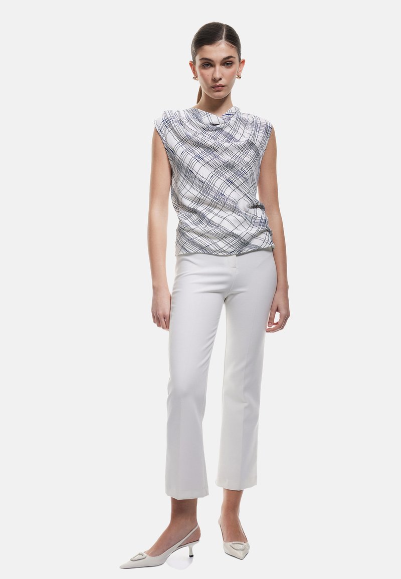Weißes ärmelloses Top mit einem blauen Karomuster, kombiniert mit weißen, enganliegenden Hosen und cremefarbenen spitzen High Heels. Glatte Textur überall.