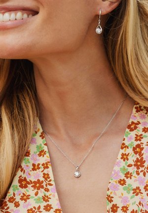Set di gioielli in argento composto da una collana con pendente a forma di goccia e orecchini abbinati con pietra trasparente. Modello che indossa un top con motivo floreale.