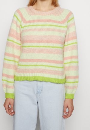 Femme portant un pull rayé pastel avec des rayures horizontales roses, blanches et vert clair et un jean bleu clair, debout devant un fond uni.