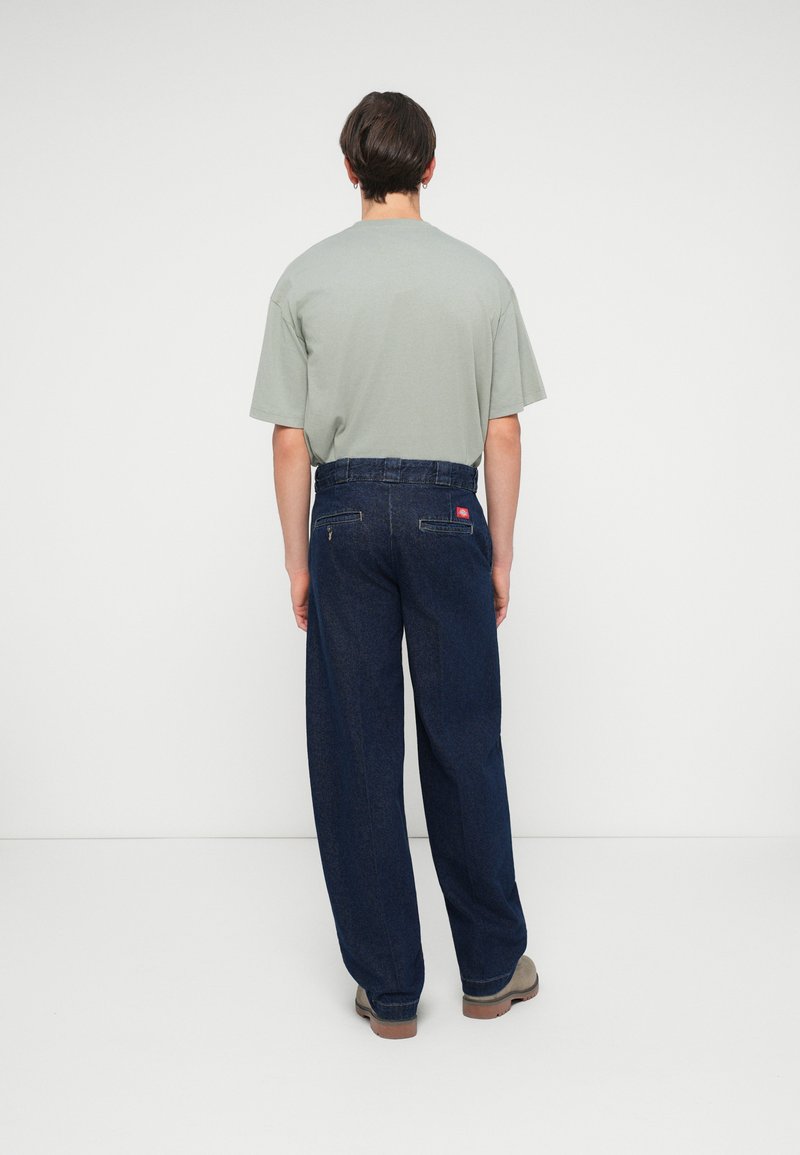 Camiseta verde clara y pantalones de mezclilla de pierna ancha azul oscuro con bolsillos traseros y etiqueta roja. El modelo lleva zapatos grises. Fondo minimalista.