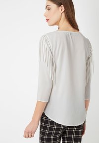 Blusa gris claro con detalles de flecos en los hombros, mangas tres cuartos y un dobladillo curvado, combinada con pantalones de cuadros en blanco y negro.