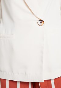 Krémové sako s jedním knoflíkem z karety, strukturovaným límcem a bočními kapsami. Stylizováno s červenobílými pruhovanými kalhotami.