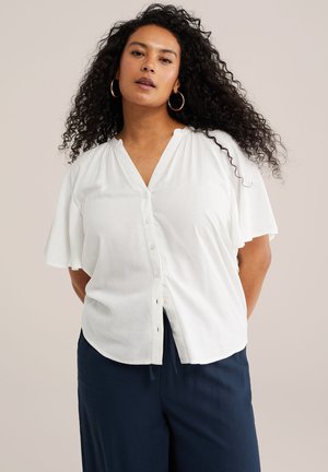 Kobieta o długich, kręconych włosach, nosząca złote kolczyki-koła, białą bluzkę zapinaną na guziki z falbaniastymi rękawami oraz granatowe spodnie, stojąca z rękami z tyłu.