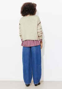 Pull beige en tricot à coupe ample, chemise rayée bordeaux en dessous, et pantalon large en denim bleu, assortis à des bottines noires.