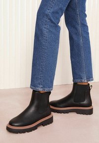 Botas de cuero negras hasta el tobillo con paneles laterales elásticos, suelas gruesas de goma y un acento marrón alrededor de la base, combinadas con jeans azules.