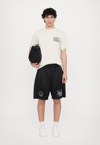 T-shirt crème clair avec impression "Aries" sur la poche, associée à un short en mesh noir avec broderie blanche. Le mannequin tient un sac noir.