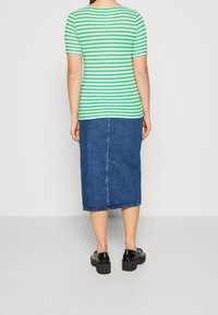 Nainen, jolla on päällään vihreä-valkopaitainen lyhythihainen paita, sininen farkkumidi-hame ja mustat platform-löysät, seisoo selin.