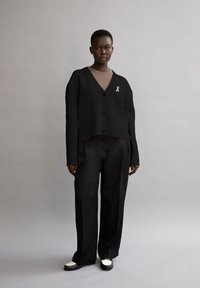 Svart kort cardigan med V-hals, knäppning fram och två framfickor, i kombination med lösa svarta byxor och tvåfärgade skor. Rosa banddetalj.
