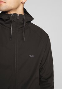 Chaqueta negra con capucha y cierre de cremallera, presenta cordones con cuentas negras y un pequeño logo "Volcom" en el pecho izquierdo.