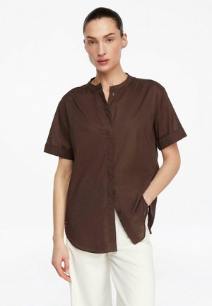 Frau trägt ein locker sitzendes dunkelbraunes Kurzarmhemd mit Knöpfen und weiße Hose, steht mit einer Hand in der Tasche vor weißem Hintergrund.