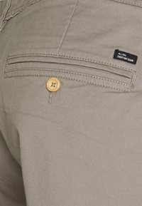 Khaki džínsové nohavice s prednou vreckom so zlatým gombíkom, prešitím a čiernou nálepkou s textom "All Day Every Day Denim."