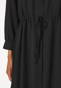 Svart kjole laget av glatt stoff, med en innsvingt midje med snøring, lange ermer og en knapp-op bodice.