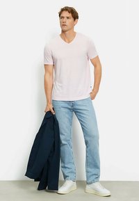 T-shirt rosa chiaro a scollo a V, in misto cotone, indossata con jeans azzurri e sneakers bianche, con in mano una giacca blu scuro. Design semplice e minimalista.