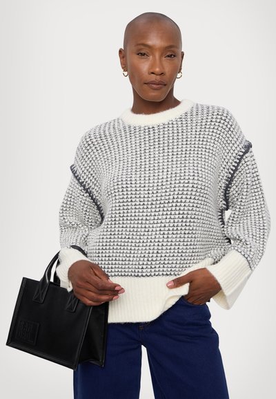 Grey and white knitted sweater with a textured pattern, featuring a ribbed white neckline and cuffs. A black leather handbag is held in hand.