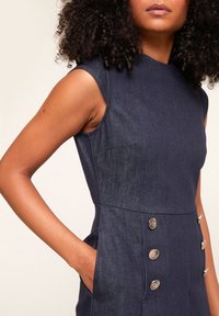 Tmavě modré džínové šaty bez rukávů, s bočními kapsami a stříbrnými kovovými knoflíky na přední straně. Hladká textura a přiléhavý střih.