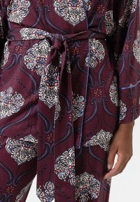 Bordeaux gekleurde wikkeljurk met bloemenpatronen in het wit, blauw en rood. Geknoopte taille en zachte stof met een gladde textuur.