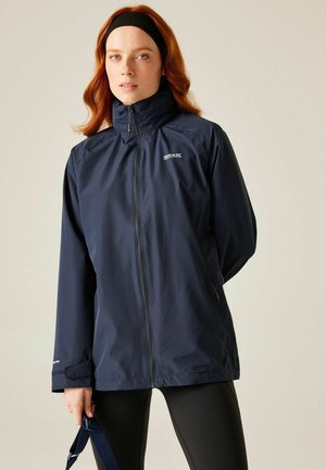 Mujer con cabello rojo que lleva una chaqueta azul marino con cremallera y leggings negros, sosteniendo una correa azul marino, de pie contra un fondo liso.