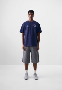 Camiseta de algodón azul marino con mangas cortas, con acentos de logo en azul, combinada con shorts de mezclilla grises y zapatillas grises, de pie contra un fondo sencillo.