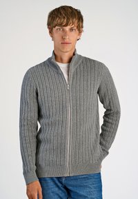 Gray knitted zip-up sweater with a ribbed texture, featuring a high collar and fitted cuffs, worn over a white t-shirt and blue jeans.