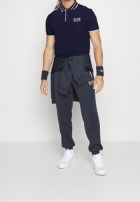 Marinblå polo-shirt med vita detaljer, i kombination med mörka träningsoveraller och vita träningsskor, med "EA7"-logotyp på båda plaggen.