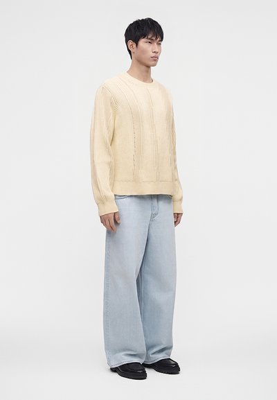Jeune homme portant un pull en maille crème, un jean large bleu clair et des chaussures noires, debout devant un fond clair uni.