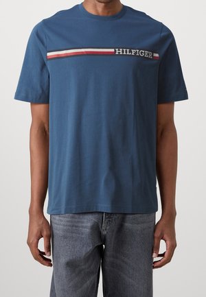 Man draagt een blauw T-shirt met een horizontale rode, witte en zwarte streep en het woord "HILFIGER" over de borst.