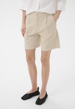 Osoba nosiaca béžové kraťasy po kolená, bielu košeľu s dlhým rukávom a čierne sieťované slip-on topánky, stojaca s rukami v vreckách.