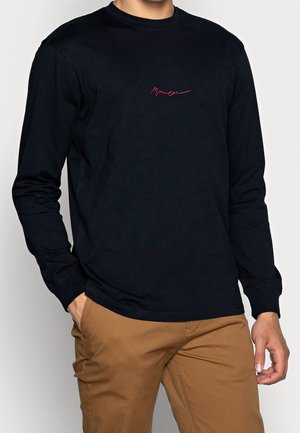 Uomo con maglia nera a maniche lunghe con logo rosso in corsivo sul petto e pantaloni marroni, mani rilassate, su uno sfondo semplice.