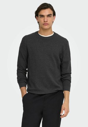Jeune homme aux cheveux foncés portant un pull gris foncé texturé par-dessus une chemise blanche et un pantalon noir, debout avec une main dans la poche.