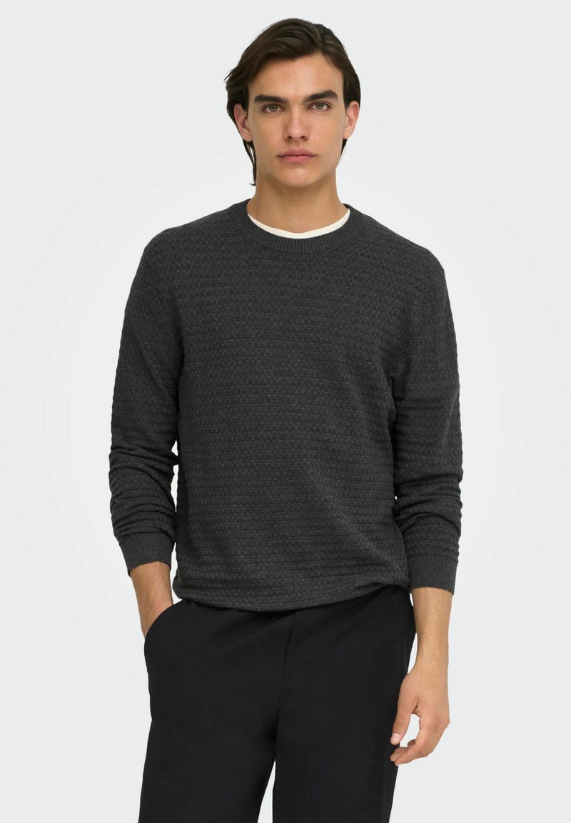 Jeune homme aux cheveux foncés portant un pull gris foncé texturé par-dessus une chemise blanche et un pantalon noir, debout avec une main dans la poche.