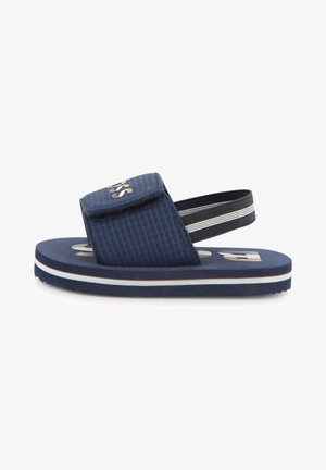 Granatowy sandał dziecięcy z regulowanym paskiem z przodu i czarnym elastycznym paskiem na pięcie, białe i brązowe paski na krawędzi podeszwy.