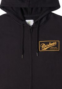 Sudadera negra con cremallera hecha de un tejido suave que presenta un parche de logo dorado en el lado izquierdo del pecho con el texto "Dcshoes SINCE 1994 WORLDWIDE."