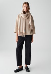 Écharpe en mélange de laine beige avec franges, portée sur un haut oversized beige clair. Un pantalon noir à jambes larges et des mocassins noirs complètent la tenue.