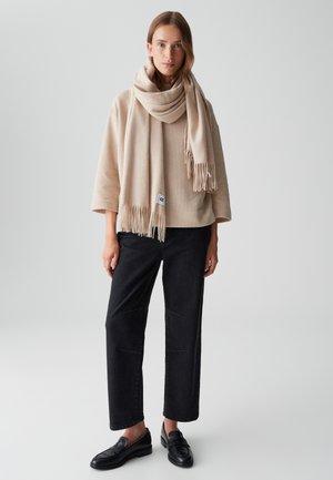 Sciarpa in misto lana beige con frange, indossata sopra un top oversized beige chiaro. I pantaloni neri a gamba larga e i mocassini neri completano l'outfit.