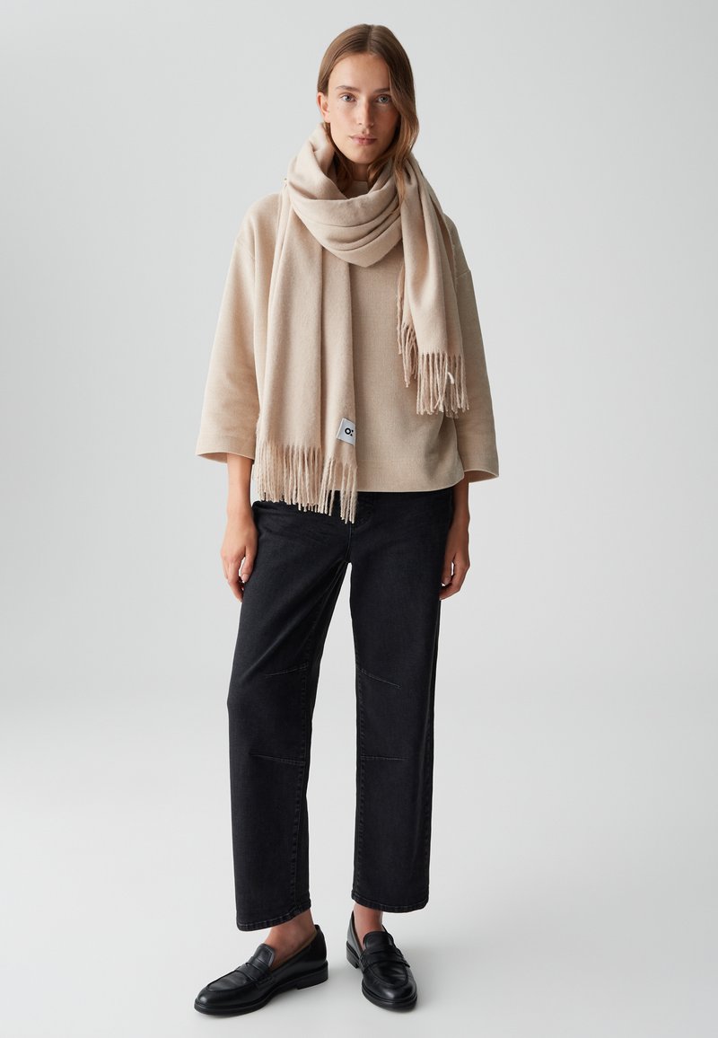 Écharpe en mélange de laine beige avec franges, portée sur un haut oversized beige clair. Un pantalon noir à jambes larges et des mocassins noirs complètent la tenue.
